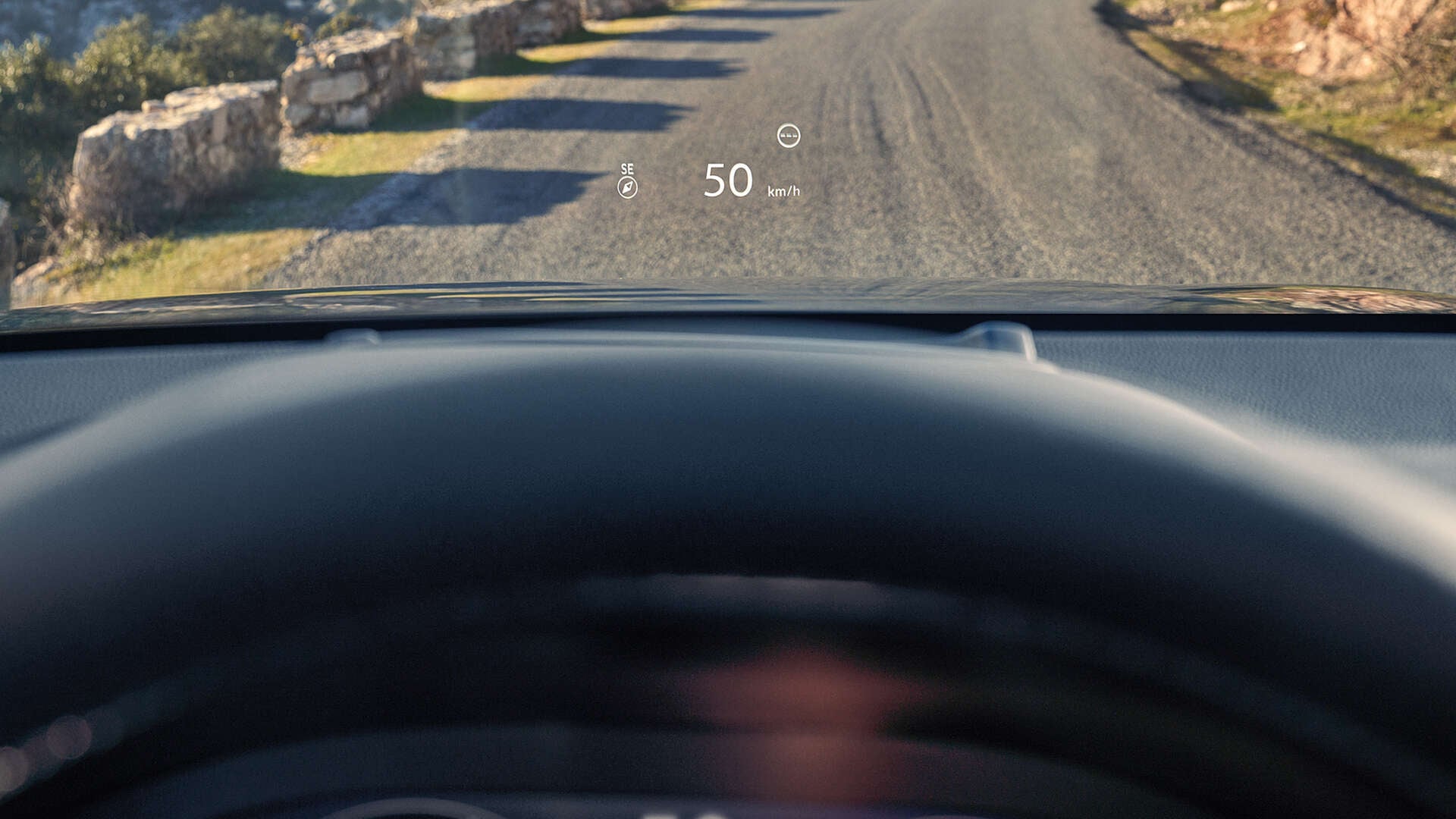 Pohľad z miesta vodiča, kde Head-Up Display (HUD) zobrazuje „50 km/h“ a kompas na čelné sklo nad štrkovou cestou.
