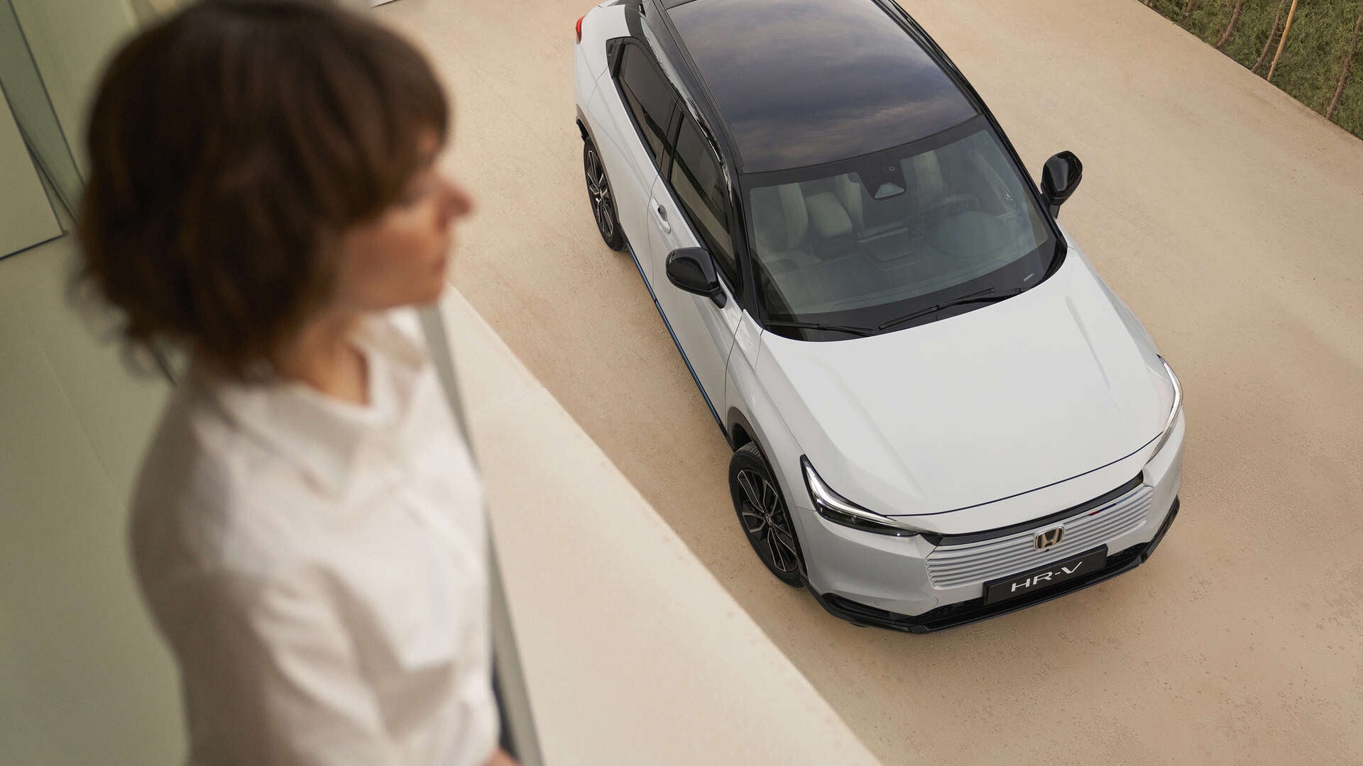 Pohľad zhora na biely Honda HR-V s čiernou strechou zaparkovaný na svetlej príjazdovej ceste, zatiaľ čo rozmazaná osoba stojí v interiéri v popredí a pozerá sa smerom k autu.